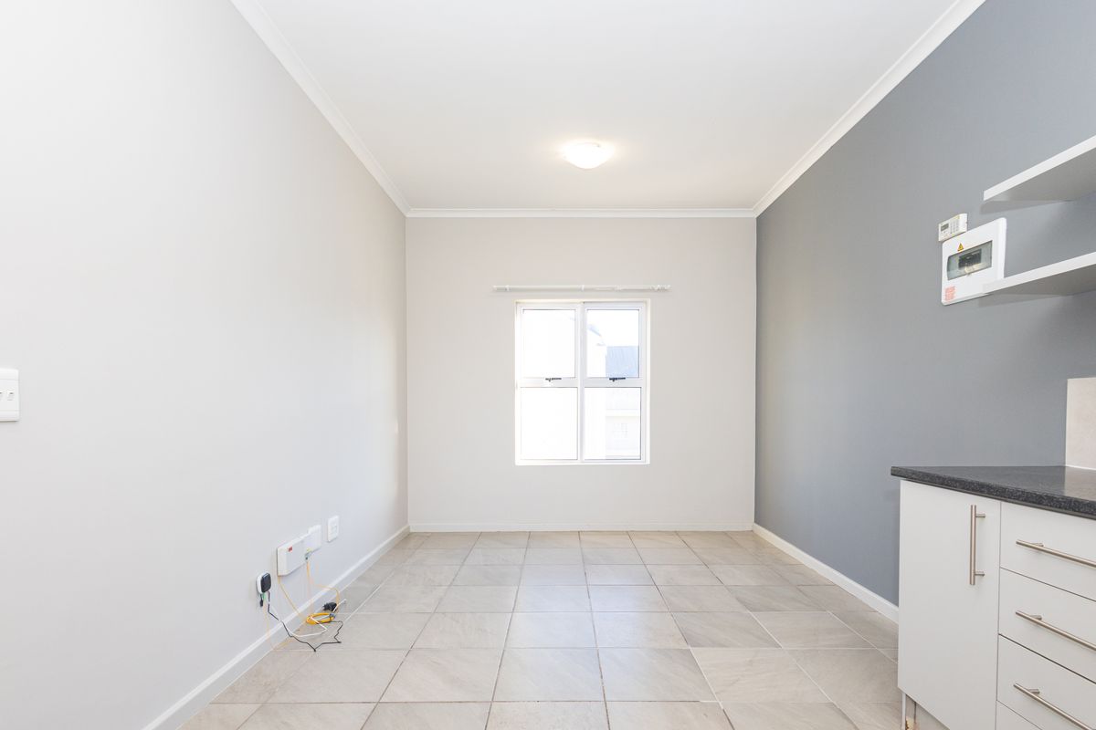 A bright, empty open-plan living area featuring neutral walls, tiled flooring, and a compact kitchen section with built-in cabinetry and a dark countertop.
