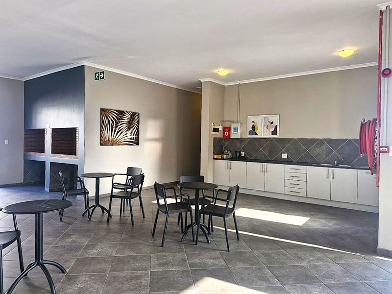 A spacious communal clubhouse area featuring a built-in braai, kitchenette with cabinetry and sink, and durable tiled flooring, suitable for social gatherings.