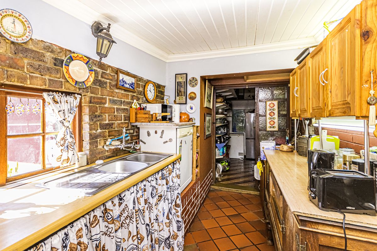 A rustic kitchen featuring exposed brick walls, terracotta flooring, timber cabinetry, and a double basin sink, leading into a secondary scullery or pantry area.