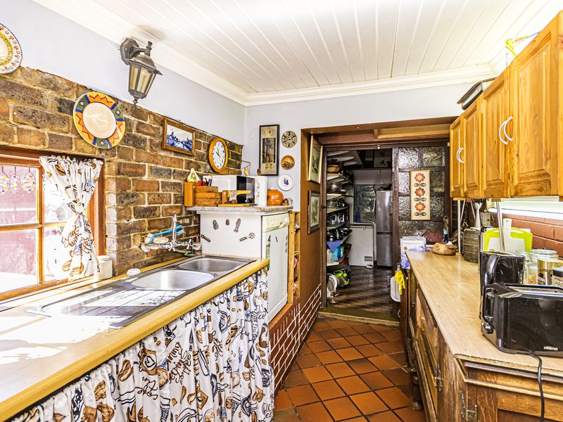 A rustic kitchen featuring exposed brick walls, terracotta flooring, timber cabinetry, and a double basin sink, leading into a secondary scullery or pantry area.