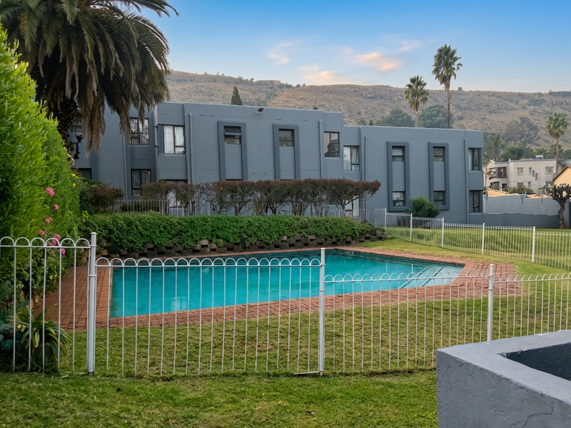 A well-maintained communal swimming pool area featuring brick coping, metal safety fencing, and landscaped garden surrounds set against a modern apartment building.