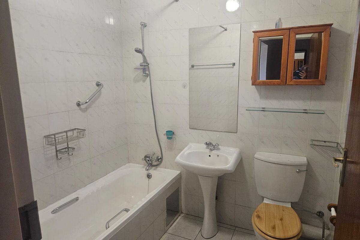 A functional bathroom featuring a bathtub with shower mixer, pedestal sink, toilet, and wall-mounted storage cabinet with tiled walls and flooring.