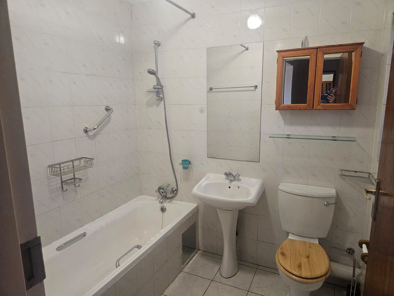 A functional bathroom featuring a bathtub with shower mixer, pedestal sink, toilet, and wall-mounted storage cabinet with tiled walls and flooring.