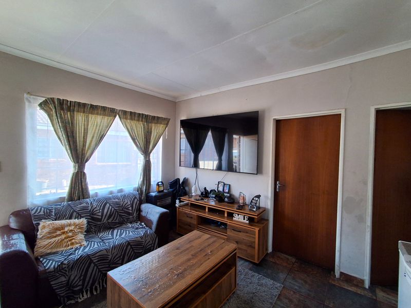 A living room featuring tiled flooring, coved ceilings, and a wall-mounted television area, showing signs of wear on the walls and ceiling.