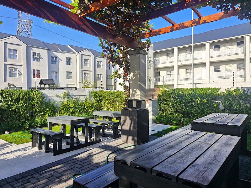 Outdoor communal patio area featuring a wooden pergola, built-in braai, and paved flooring, surrounded by established garden hedging within an apartment complex.