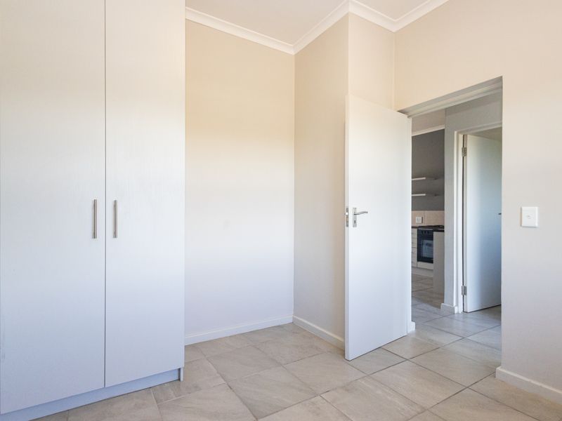 A bright, empty bedroom featuring neutral walls, tiled flooring, and a built-in wardrobe, with a view into the adjacent kitchen area.