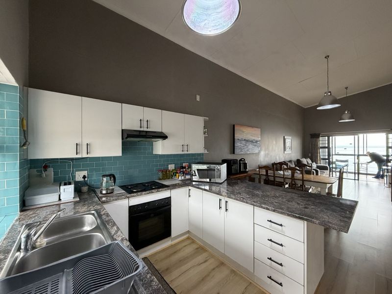 Open-plan kitchen featuring white cabinetry, dark laminate countertops, a tiled backsplash, and integrated appliances, flowing into the dining and living area.