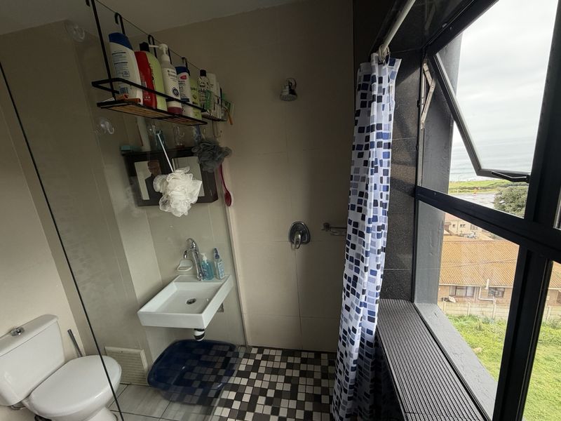 A functional bathroom featuring a wall-mounted basin, shower area, and toilet, complemented by a large window offering natural light and coastal views.
