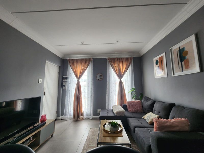 A well-maintained living room featuring neutral grey walls, tiled flooring, and ample natural light through double windows with elegant drapery.