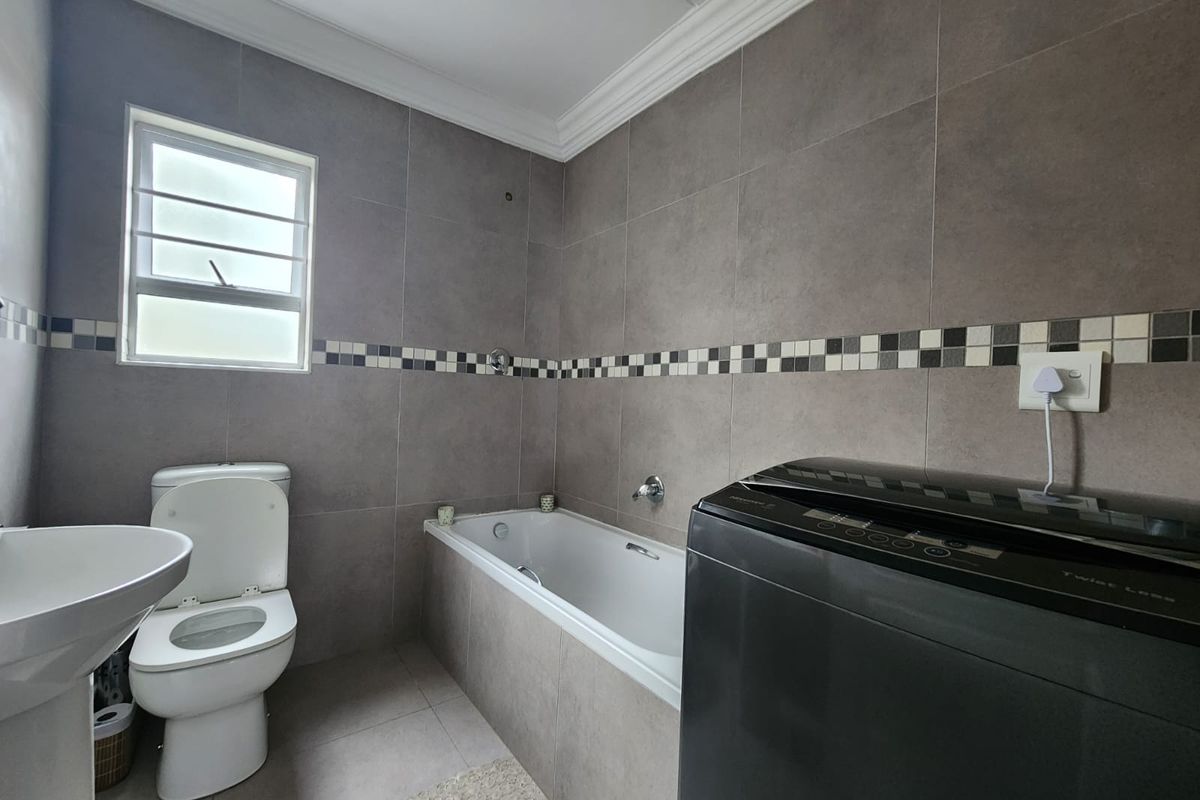 A modern bathroom featuring neutral grey wall and floor tiles, a built-in bathtub, a pedestal basin, and a toilet.