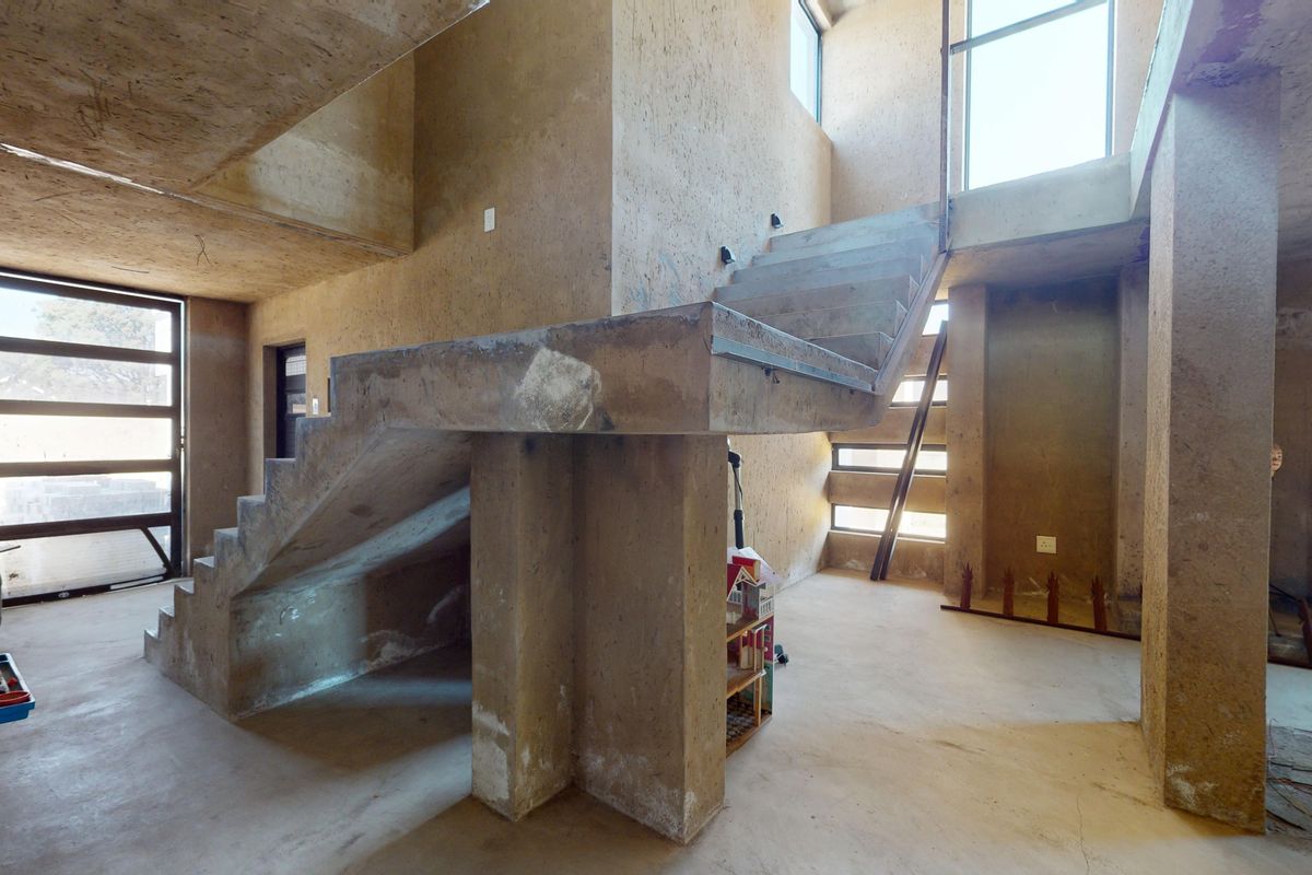 An open-plan living area featuring raw exposed concrete walls, a central concrete staircase, and large windows providing natural light in an unfinished state.