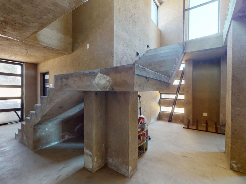 An open-plan living area featuring raw exposed concrete walls, a central concrete staircase, and large windows providing natural light in an unfinished state.