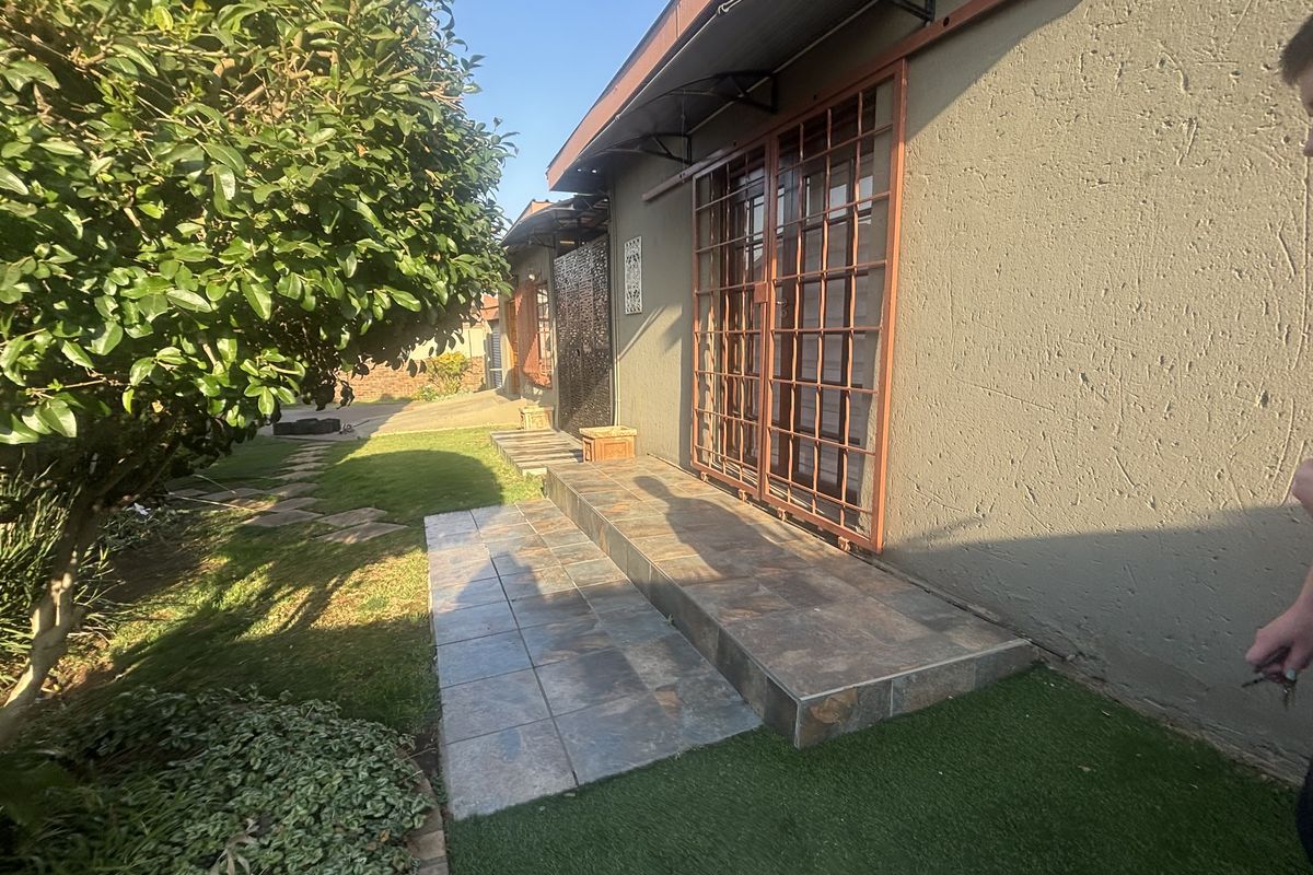 Rear exterior view featuring a tiled patio area, sliding door with security gate, and an awning, set against a landscaped garden.