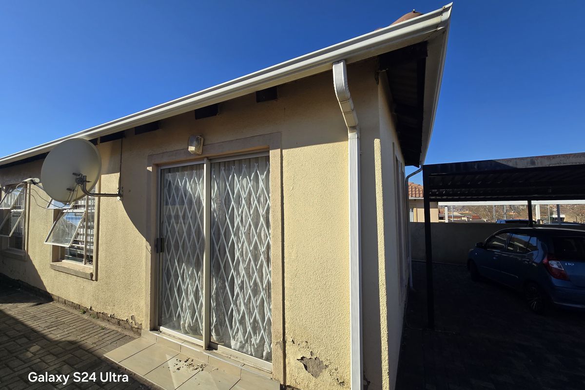 Rear exterior view of the property featuring a sliding door with security gate, paved patio area, and adjacent carport structure.