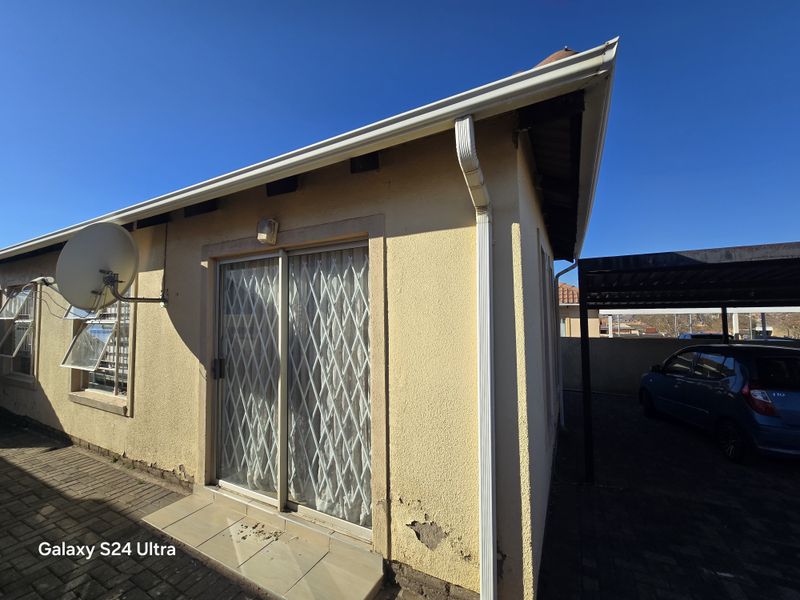 Rear exterior view of the property featuring a sliding door with security gate, paved patio area, and adjacent carport structure.
