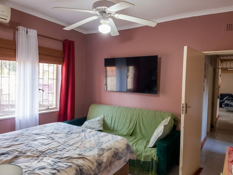 A bedroom featuring a ceiling fan, wall-mounted air conditioning unit, and security bars on the window, with a view into the adjacent hallway.