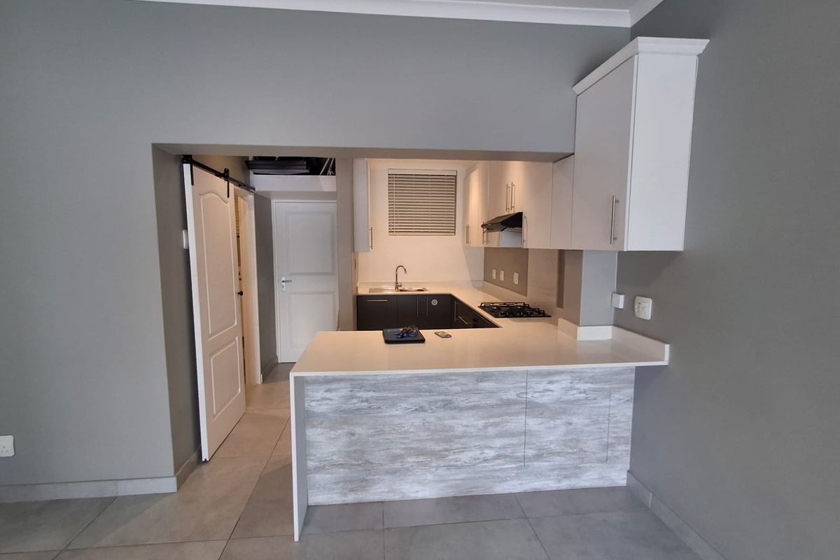 A modern, compact kitchen featuring white cabinetry, stone countertops, a gas hob, and an integrated breakfast bar, finished with neutral grey walls and tiled floors.