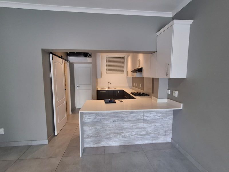 A modern, compact kitchen featuring white cabinetry, stone countertops, a gas hob, and an integrated breakfast bar, finished with neutral grey walls and tiled floors.