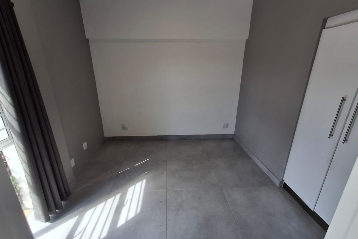A bright, empty bedroom featuring modern grey floor tiles, neutral wall tones, and a built-in wardrobe with sleek handles.