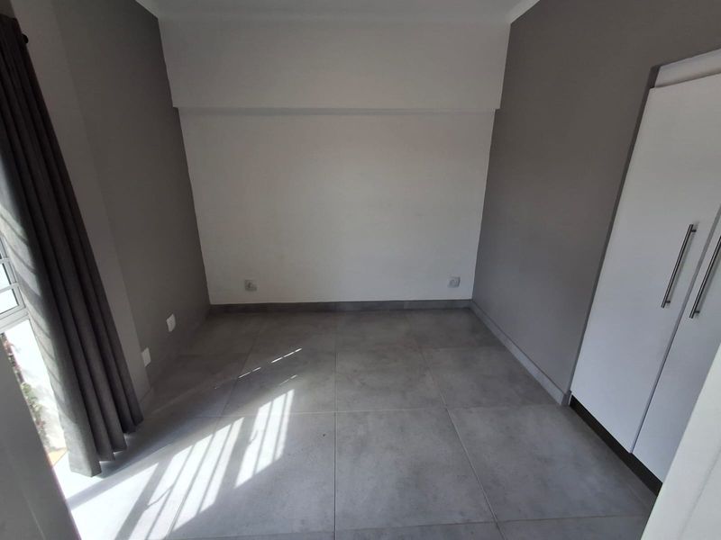 A bright, empty bedroom featuring modern grey floor tiles, neutral wall tones, and a built-in wardrobe with sleek handles.