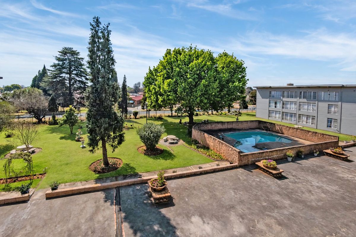 Aerial view of the apartment complex grounds featuring a swimming pool, expansive lawn, mature trees, and a paved outdoor terrace area.