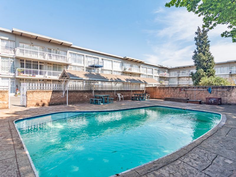 Communal swimming pool area featuring a paved deck, boundary wall, and a covered seating area within an apartment complex setting.