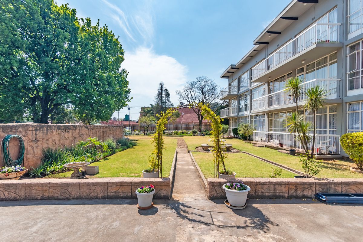 A well-maintained rear garden area featuring a paved walkway, decorative metal arch, and a multi-story apartment building with private balconies.