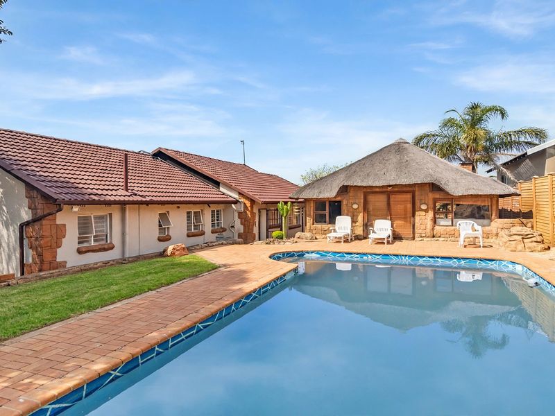 Outdoor pool area featuring a swimming pool, brick-paved surround, and a traditional thatched lapa, set within a well-maintained garden environment.