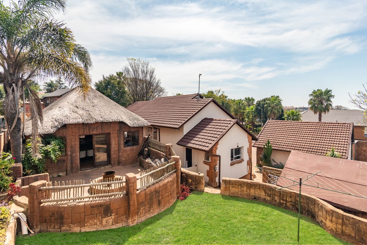 Rear view of the property featuring a thatched roof lapa, tiled roof main structure, stone-clad retaining walls, and a well-maintained garden area.