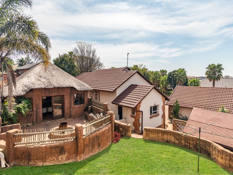 Rear view of the property featuring a thatched roof lapa, tiled roof main structure, stone-clad retaining walls, and a well-maintained garden area.
