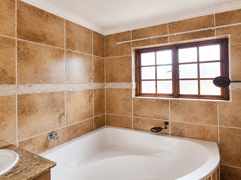 A bathroom featuring a corner bathtub, tiled walls, and a vanity countertop with a drop-in sink, illuminated by a wooden-framed window.