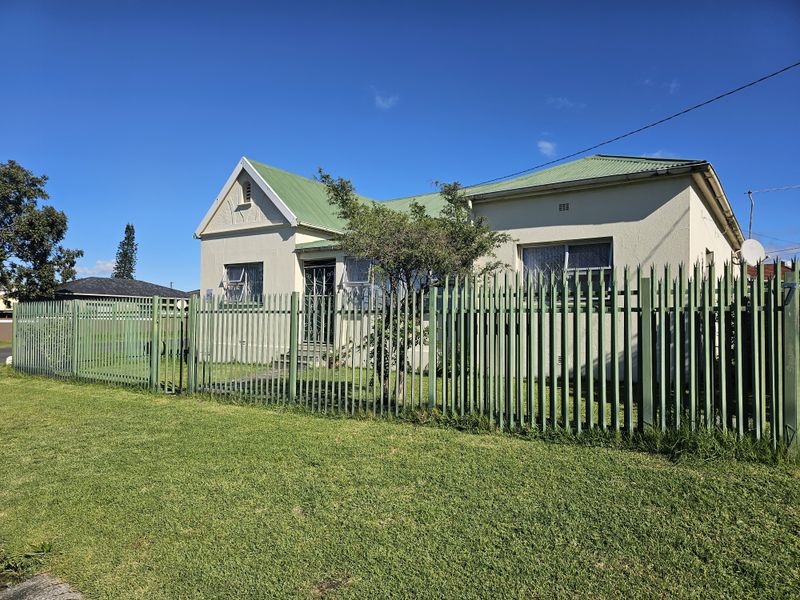 Front view of a single-story townhouse featuring a green corrugated metal roof, plastered exterior walls, and secure perimeter fencing with a gated entrance.