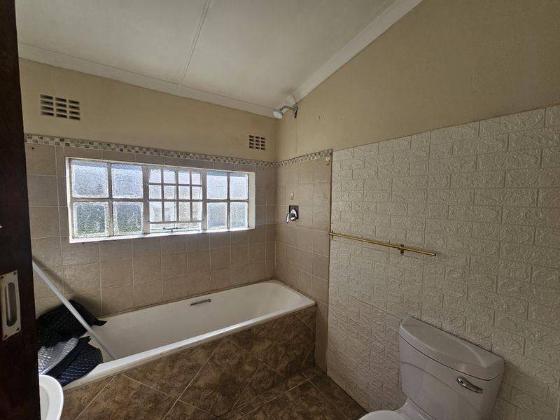 A dated bathroom featuring a built-in bathtub, shower head, and toilet, with tiled wall accents and a window for natural light.