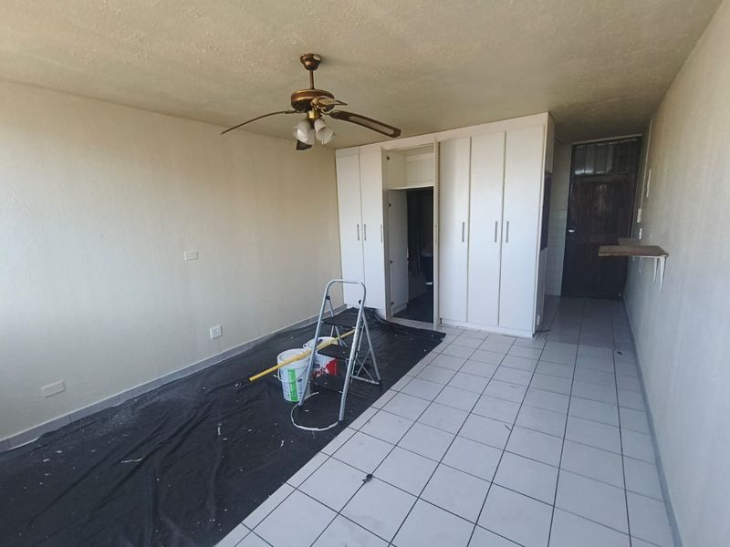A vacant bedroom featuring built-in cupboards, tiled flooring, and a ceiling fan, currently undergoing minor maintenance and painting.