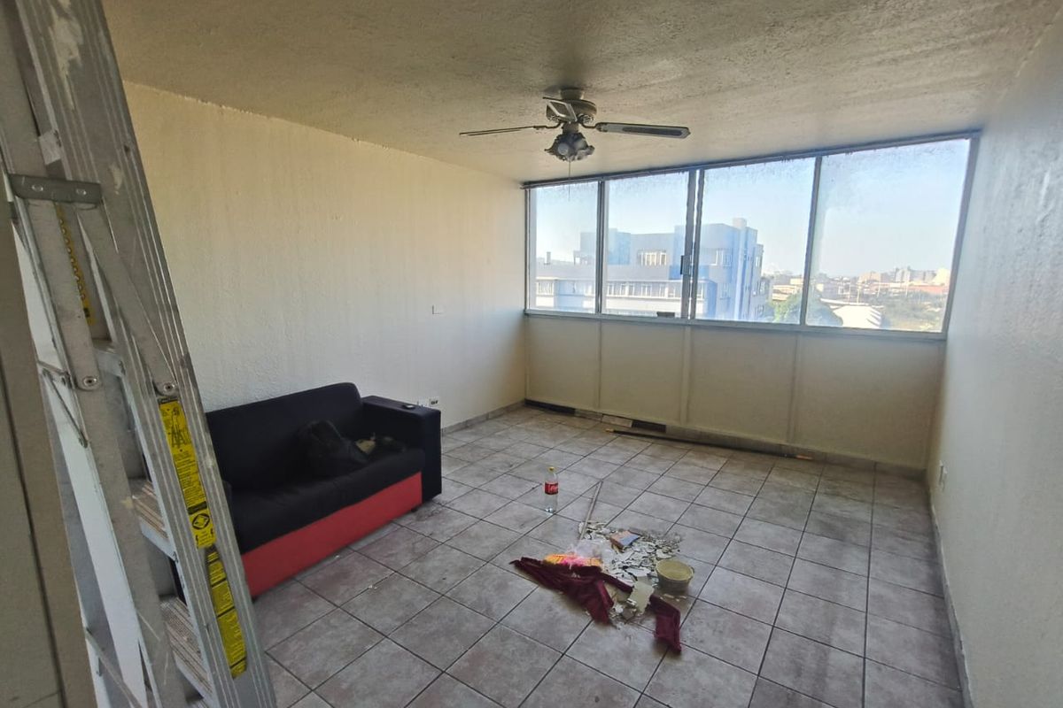 A sunlit living area featuring tiled flooring, a ceiling fan, and large windows with an urban view, currently in need of renovation.