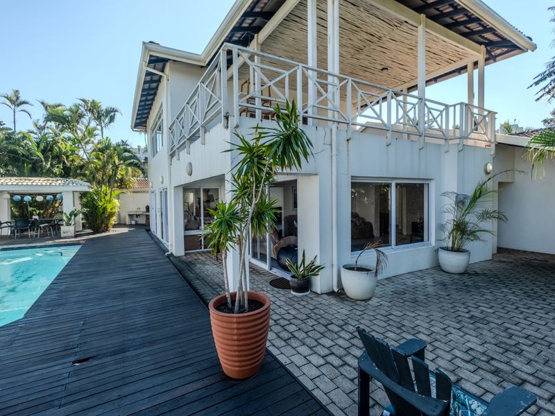 Rear exterior view featuring a swimming pool, timber decking, paved patio area, and a covered upper-level balcony with sliding glass door access.