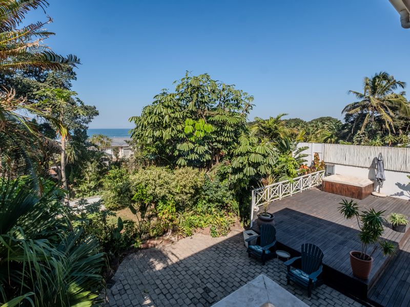 Rear exterior view featuring a timber deck, paved patio area, built-in hot tub, and lush garden with distant ocean views.