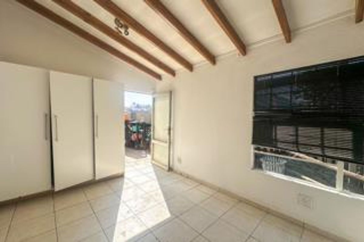 A bright, empty bedroom featuring tiled flooring, exposed timber ceiling beams, and built-in cupboards, with direct access to an outdoor area.