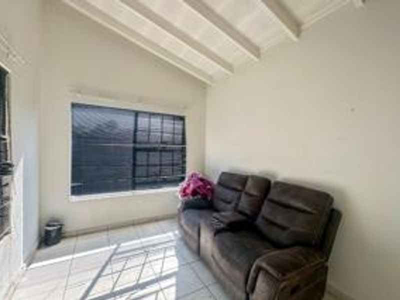 A living room featuring tiled flooring, exposed beam ceilings, and large windows with fitted venetian blinds.