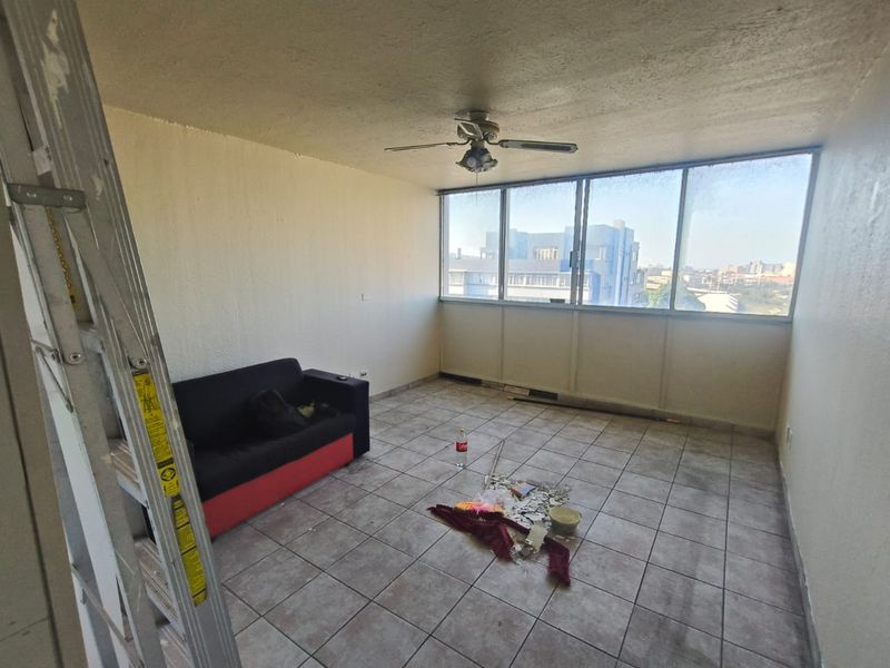 A sunlit living area featuring tiled flooring, a ceiling fan, and large windows with an urban view, currently in need of renovation.