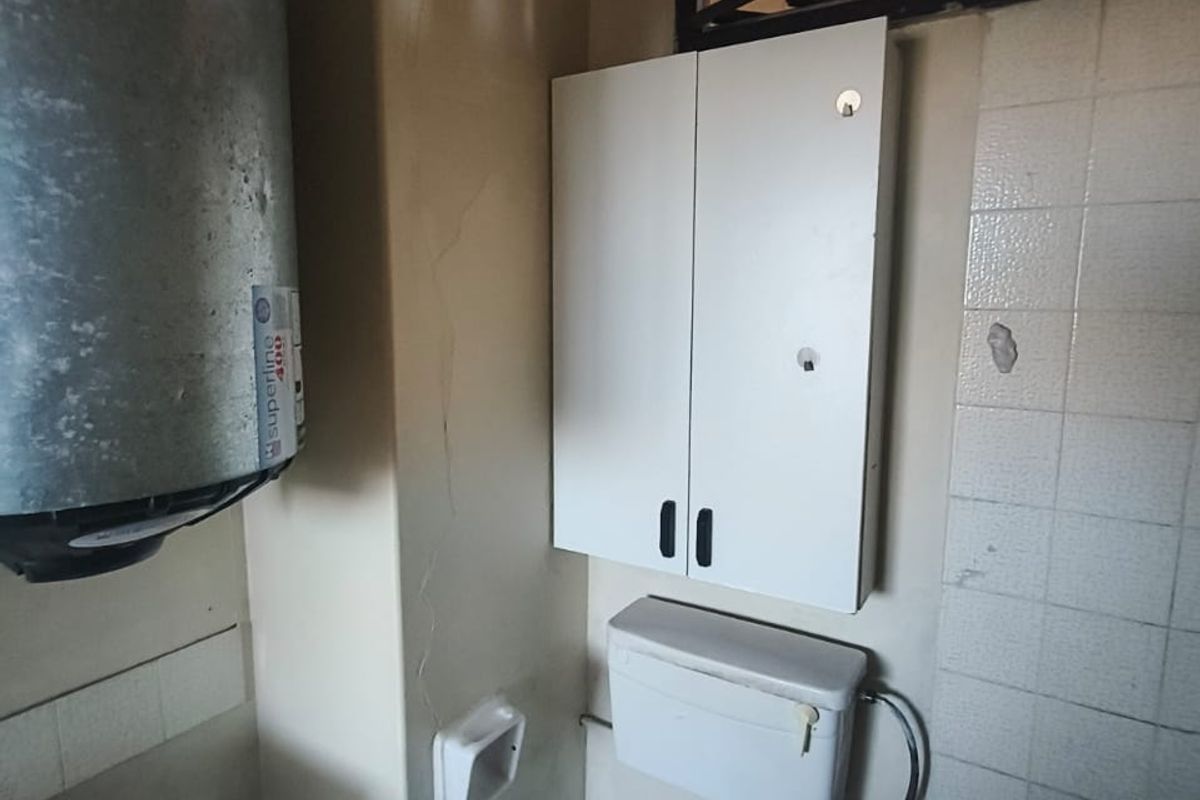 A dated bathroom featuring an exposed electric water heater, wall-mounted storage, and basic sanitary fixtures, requiring significant renovation and cosmetic updates.