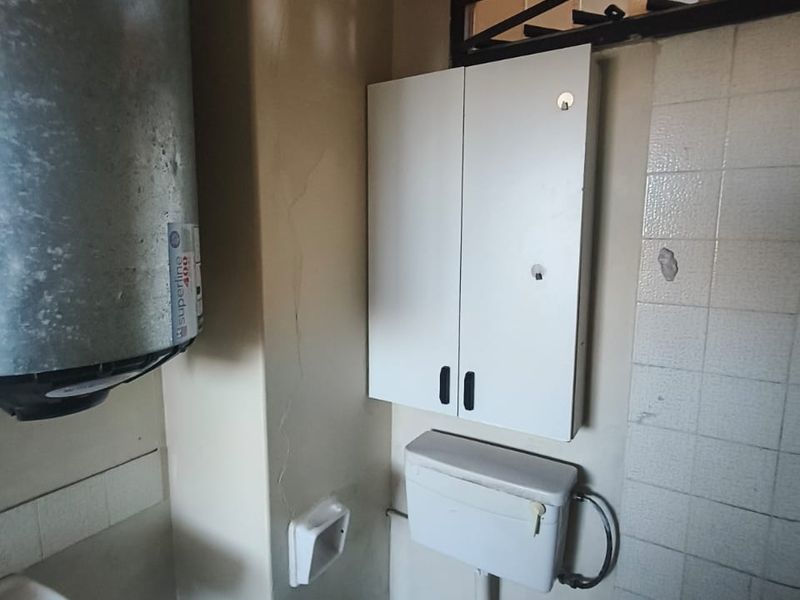 A dated bathroom featuring an exposed electric water heater, wall-mounted storage, and basic sanitary fixtures, requiring significant renovation and cosmetic updates.