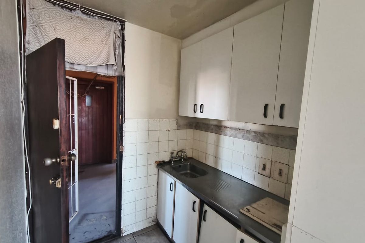 A compact, dated kitchen area featuring basic built-in cabinetry, a stainless steel sink, and tiled backsplash, requiring significant renovation and cosmetic updates.