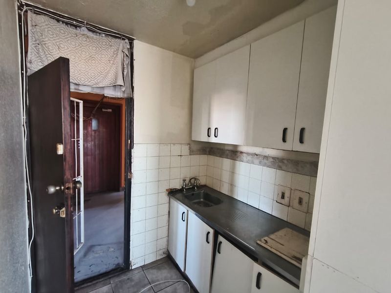 A compact, dated kitchen area featuring basic built-in cabinetry, a stainless steel sink, and tiled backsplash, requiring significant renovation and cosmetic updates.