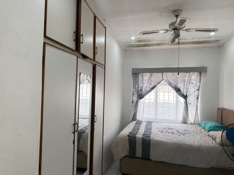A bedroom featuring built-in cupboards, a ceiling fan, and a window with security bars. The ceiling shows visible signs of water damage.
