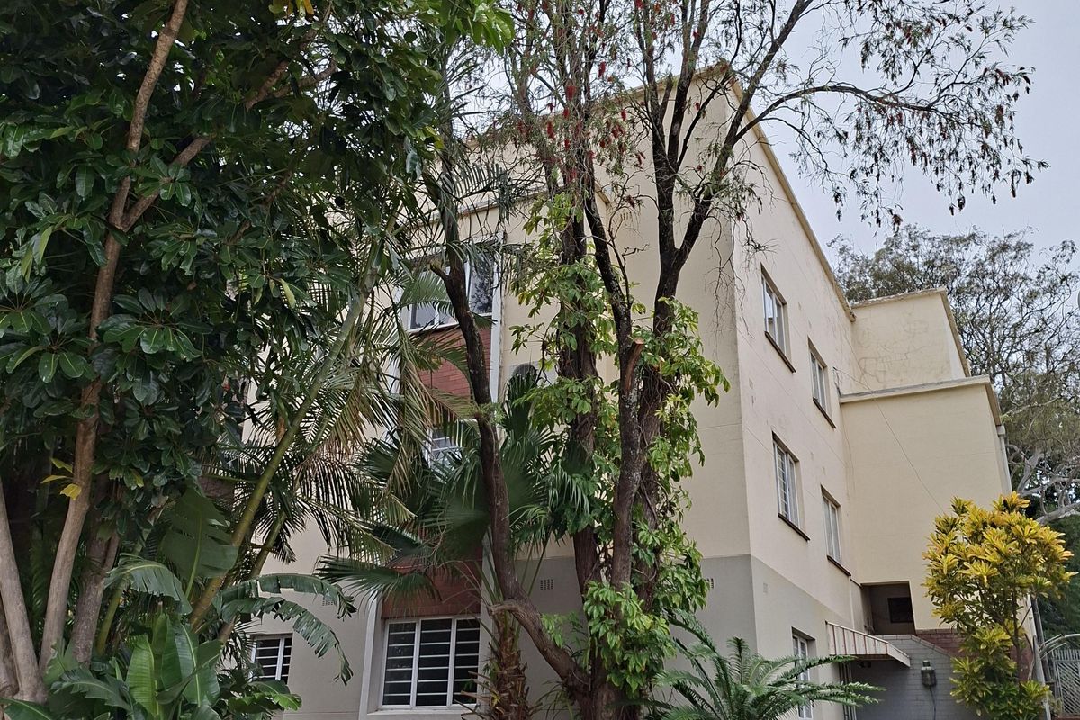 Exterior view of a multi-story apartment building featuring a landscaped garden, paved walkway, and security-barred windows.