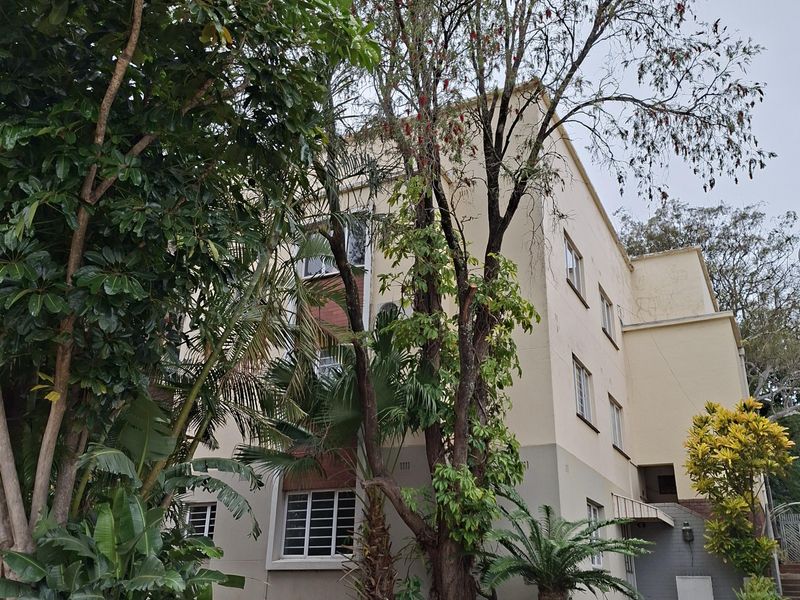 Exterior view of a multi-story apartment building featuring a landscaped garden, paved walkway, and security-barred windows.