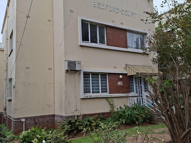 Front elevation of Bedford Court apartment building featuring a plastered facade with brick accents, metal window frames, and an external air conditioning unit.