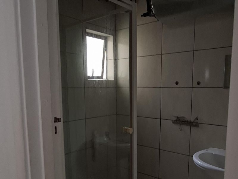A small, dated bathroom featuring a pedestal basin and a shower enclosure with tiled walls and floor, requiring modernization and improved lighting.
