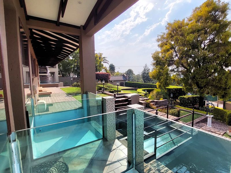 A luxurious outdoor entertainment area featuring a modern glass-fronted pool, covered patio, and beautifully landscaped tiered gardens.